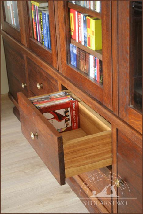 Dębowa biblioteczka z przeszklonymi półkami i wysuwaną szufladą wypełnioną płytami CD, widoczny detal wykonania szuflady z jasnego drewna.