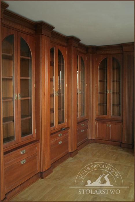 Drewniana biblioteka z rzeźbionymi detalami, przeszklonymi szafkami i szufladami, tworząca narożną zabudowę, widoczny parkiet na podłodze oraz logo stolarza na podłodze.