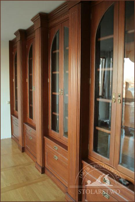Rząd wysokich, fornirowanych biblioteczek z przeszklonymi drzwiami i szufladami, w stylu klasycznym, z widocznym logo stolarza w prawym dolnym rogu.
