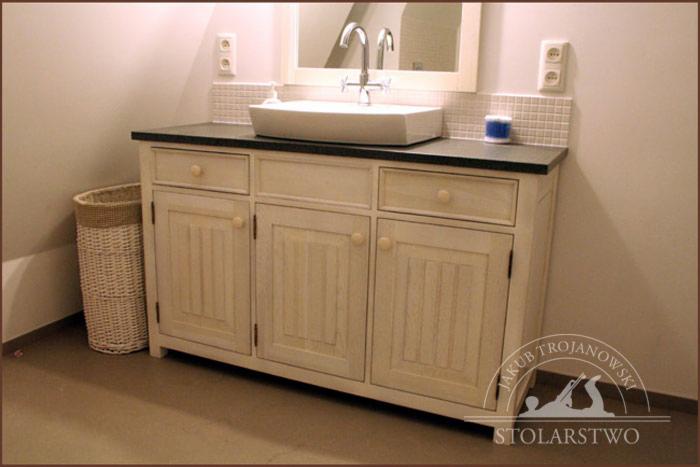 Biała komoda łazienkowa z trzema szufladami i szafkami, czarnym blatem i nablatową umywalką, uzupełniona wiklinowym koszem w rogu pomieszczenia.