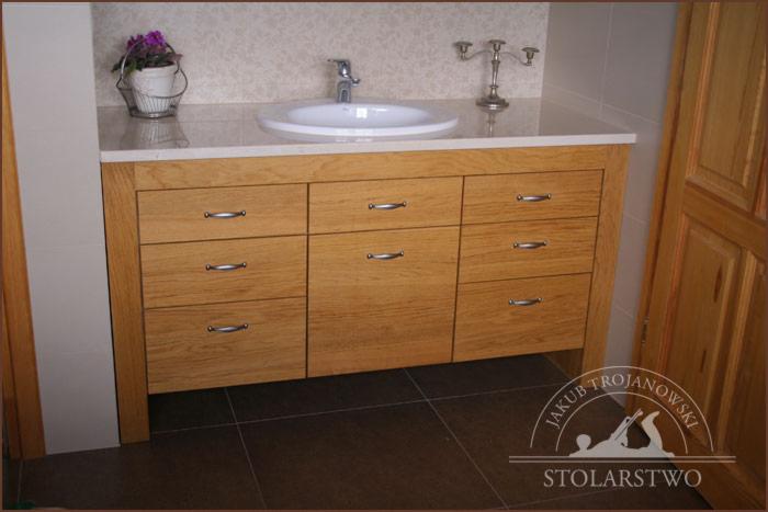 Drewniana komoda łazienkowa z dziewięcioma szufladami i chromowanymi uchwytami, umywalka nablatowa i bateria, na blacie świecznik i doniczka z kwiatami, widoczne logo firmy.