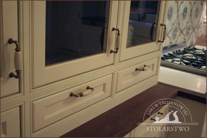 Fragment kremowej zabudowy kuchennej w stylu angielskim z przeszklonymi szafkami i ceramicznymi uchwytami w kolorze ecru, widoczny blat z ciemnego drewna oraz logo stolarza.