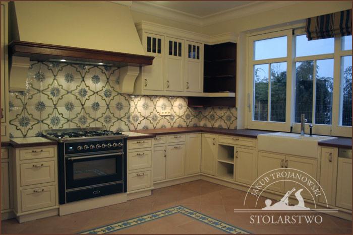 Kremowa kuchnia w stylu angielskim z ciemnymi drewnianymi blatami, wzorzystymi płytkami nad kuchenką i dużym oknem z widokiem na zewnątrz.