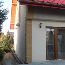 Elewacja domu z oknami i lampą zewnętrzną, wykończona w jasnych kolorach z dekoracyjnym, piaskowym elementem narożnym. Czerwony dach i zieleń drzew w tle.