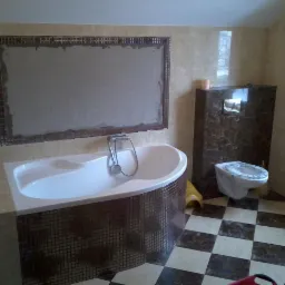 Łazienka w trakcie wykończenia: wanna obudowana mozaiką, toaleta i ściany wyłożone płytkami. Na ścianie przygotowane miejsce pod lustro z mozaikową ramą.