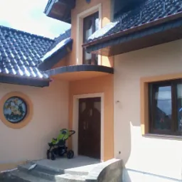 Wykończona elewacja domu w Popowie z okrągłym oknem i wózkiem dziecięcym przy wejściu. Widoczne rusztowanie po prawej stronie, sugerujące niedawne prace.