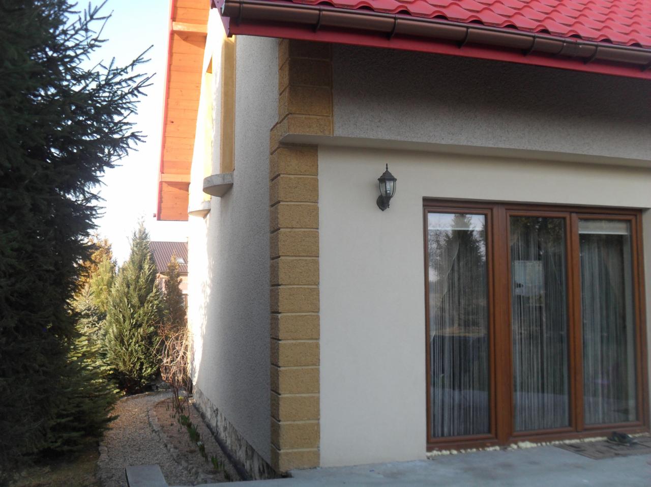 Elewacja domu z oknami i lampą zewnętrzną, wykończona w jasnych kolorach z dekoracyjnym, piaskowym elementem narożnym. Czerwony dach i zieleń drzew w tle.