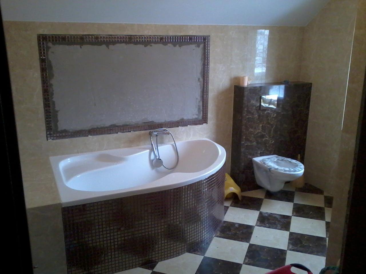 Łazienka w trakcie wykończenia: wanna obudowana mozaiką, toaleta i ściany wyłożone płytkami. Na ścianie przygotowane miejsce pod lustro z mozaikową ramą.