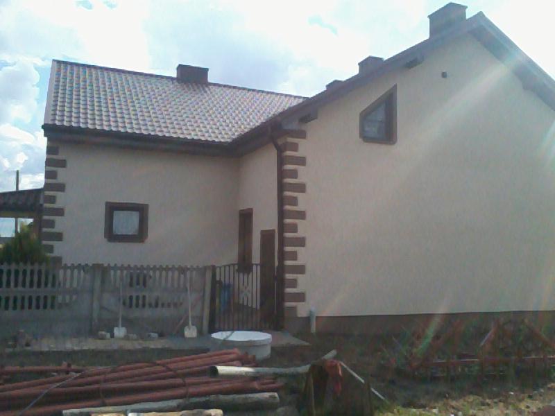 Dom jednorodzinny z beżową elewacją i brązowymi elementami dekoracyjnymi wokół okien i na rogach, widziany z boku w słoneczny dzień.