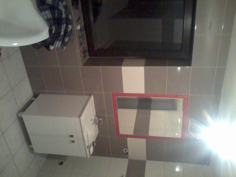 Wykończona łazienka z mozaiką: umywalka z szafką, lustro w czerwonej ramie z mozaiki, okno i płytki w odcieniach szarości i bieli. Ujęcie z góry.