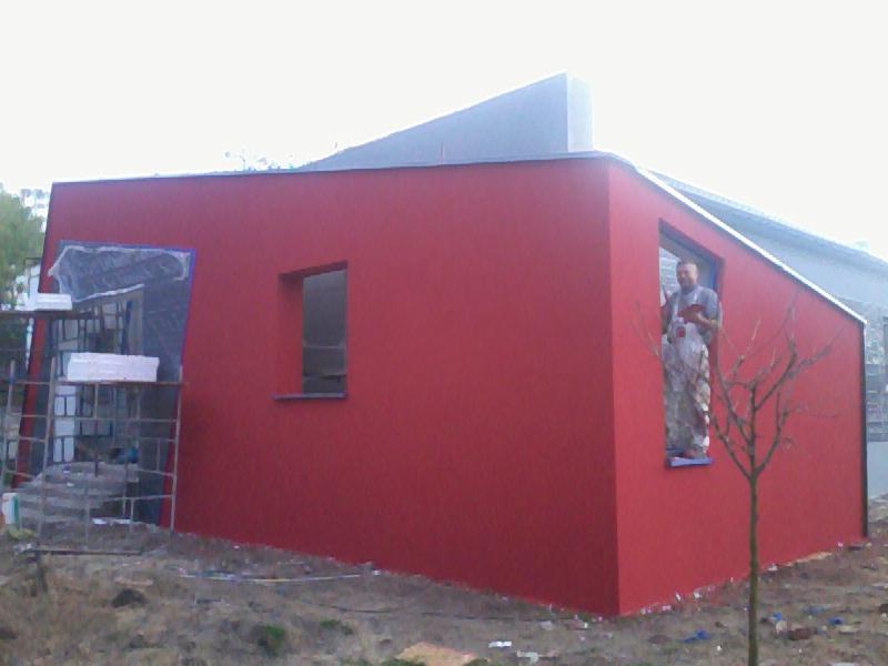 Czerwona elewacja budynku z oknami, malowana przez pracownika w kombinezonie. Część okien zabezpieczona folią, widoczne rusztowania i drzewko.