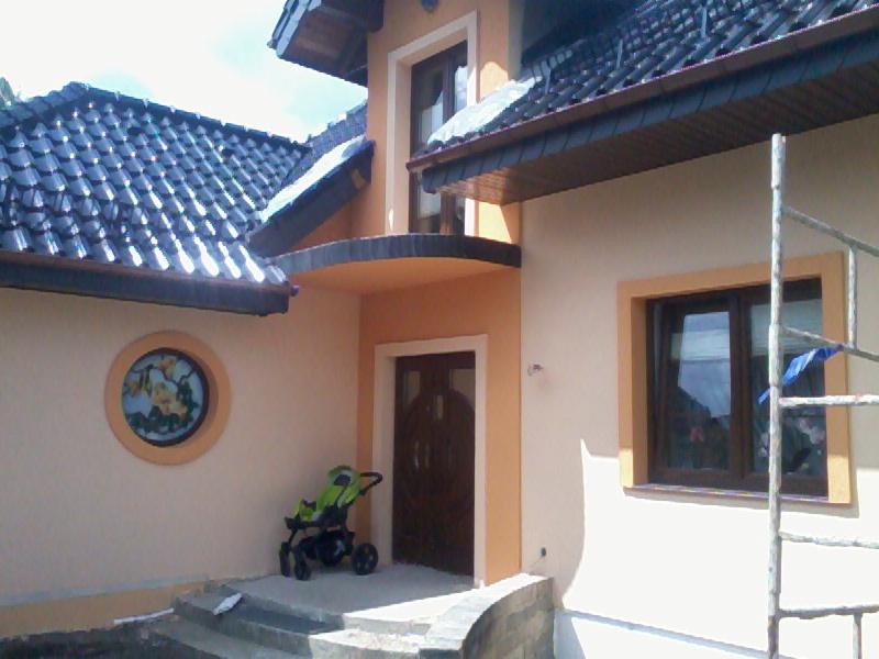 Wykończona elewacja domu w Popowie z okrągłym oknem i wózkiem dziecięcym przy wejściu. Widoczne rusztowanie po prawej stronie, sugerujące niedawne prace.