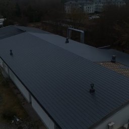 ANETA STEFANIAK SEBA-DACHY USŁUGI CIESIELSKO DEKARSKO BUDOWLANE - Ciemnoszary dach budynku z blachy trapezowej, częściowo w trakcie wymiany. Widoczne kominy i elementy konstrukcji dachu. Ujęcie z góry, pochmurny dzień.