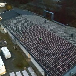 ANETA STEFANIAK SEBA-DACHY USŁUGI CIESIELSKO DEKARSKO BUDOWLANE - Wieczorny widok z góry na duży budynek z częściowo wymienionym dachem. Widoczna konstrukcja dachu, pracownicy w odblaskowych kamizelkach i zaparkowane pojazdy.