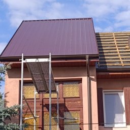 ANETA STEFANIAK SEBA-DACHY USŁUGI CIESIELSKO DEKARSKO BUDOWLANE - Częściowo pokryty, bordowy dach z blachy trapezowej na budynku. Widoczne rusztowanie i fragment dachu z łatami. Błękitne niebo w tle.