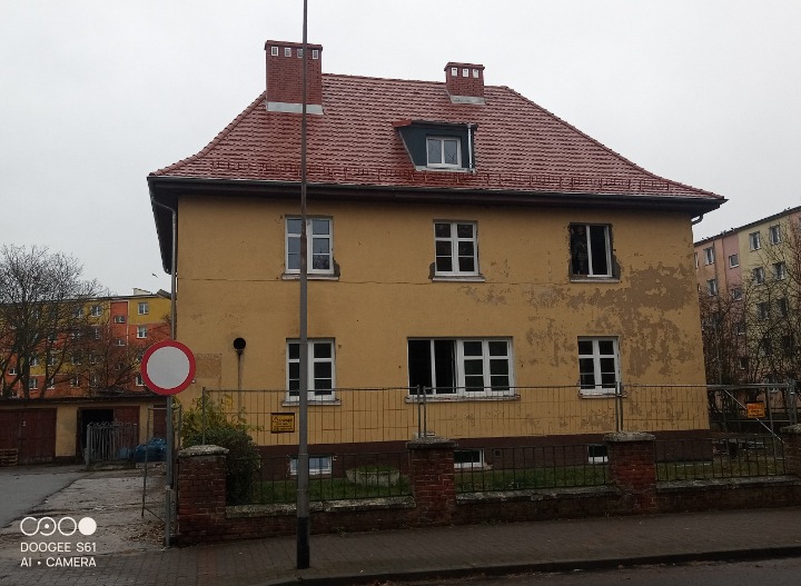 Dwupiętrowy budynek z czerwonym dachem z cegły i dwoma kominami, z żółtą elewacją z widocznymi śladami uszkodzeń, ogrodzony metalowym płotem, z widocznym znakiem zakazu w tle.