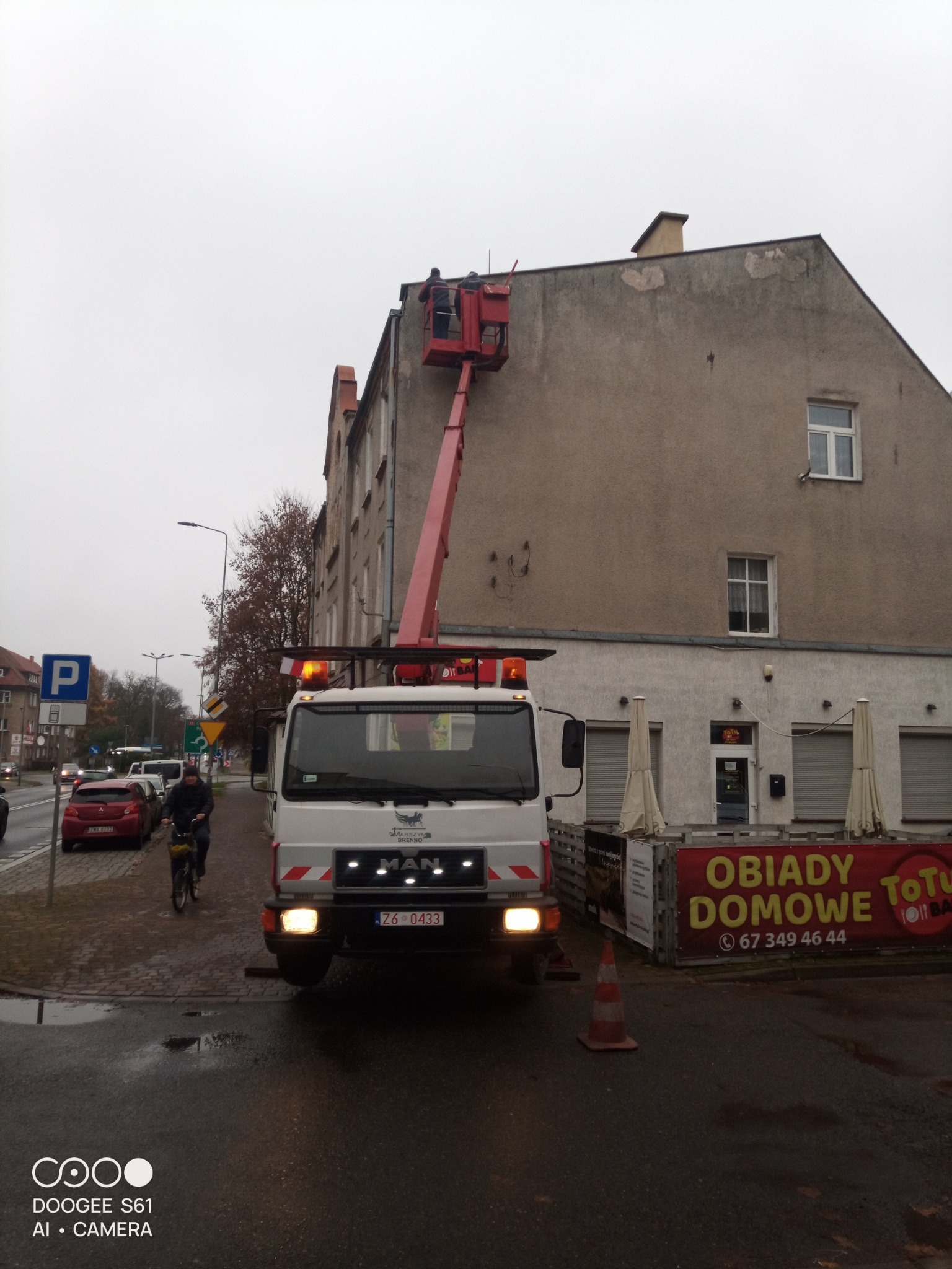 Praca na wysokościach z użyciem podnośnika koszowego przy budynku. Widoczny pracownik w koszu, pojazd MAN z włączonymi światłami ostrzegawczymi i rowerzysta na chodniku.