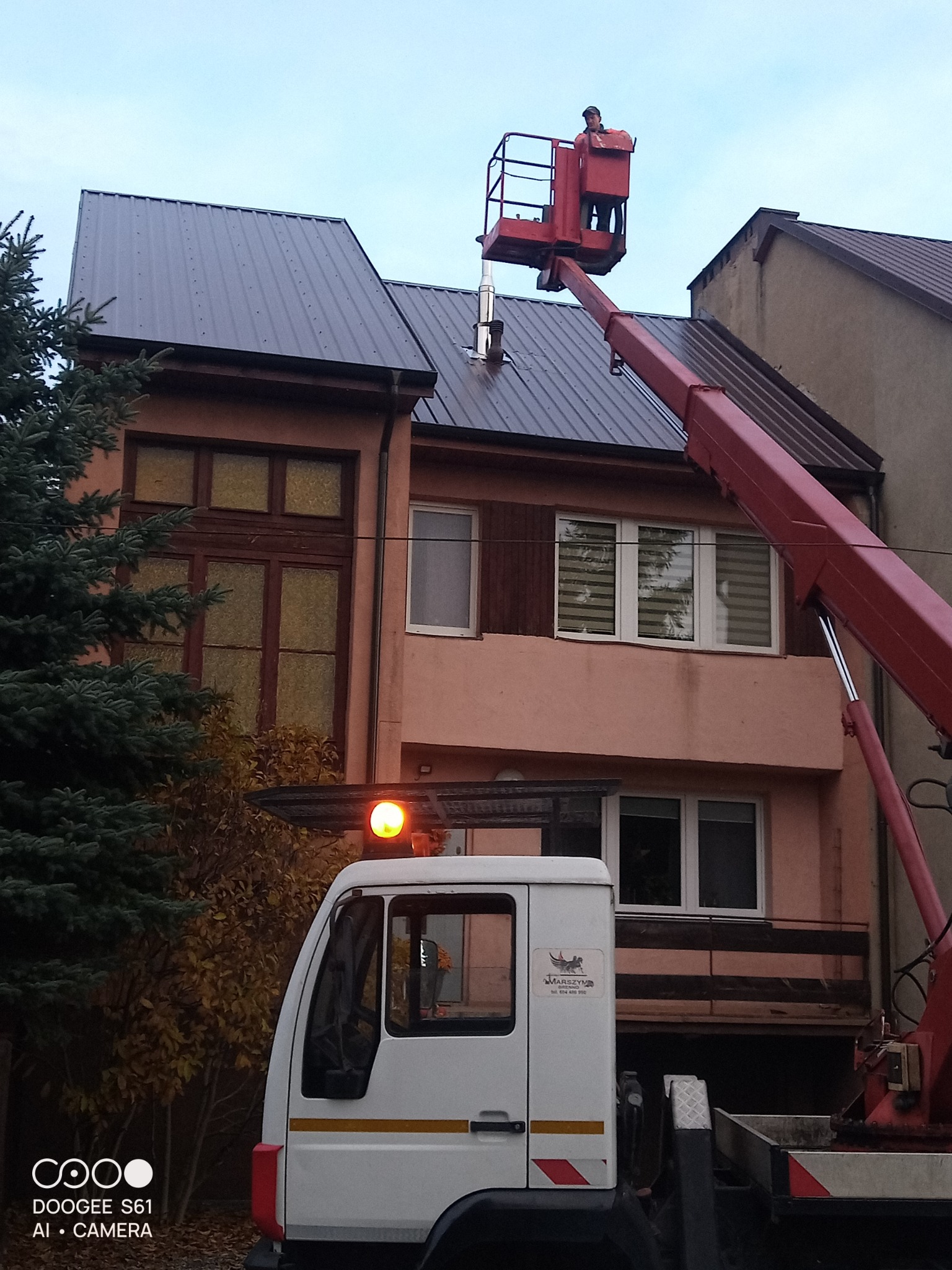 Pracownik na podnośniku koszowym przy dachu domu z blachy trapezowej. Widoczny fragment elewacji i drzewo iglaste. Praca na wysokości.