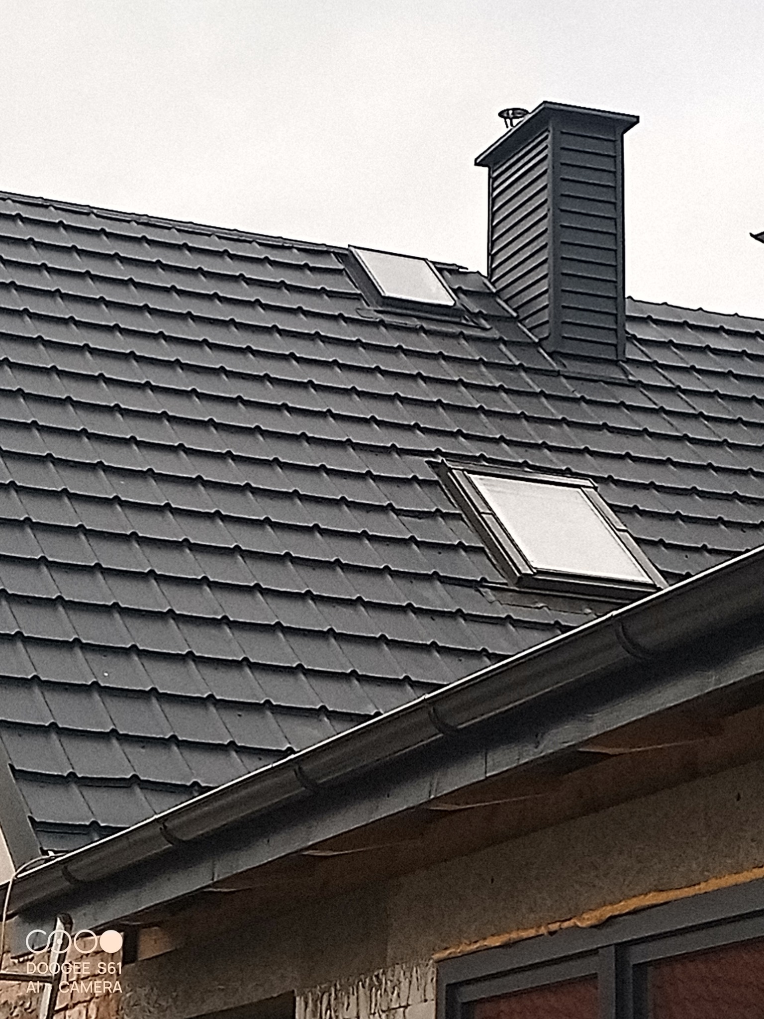Czarny dach z dachówki modułowej, dwa okna dachowe i komin. Widoczne rynny i fragment ściany budynku. Ujęcie z dołu, pochmurne niebo.