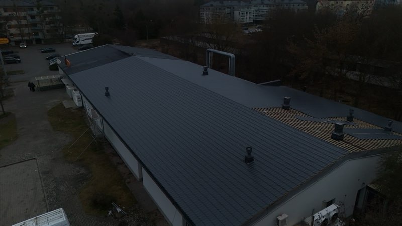 Ciemnoszary dach budynku z blachy trapezowej, częściowo w trakcie wymiany. Widoczne kominy i elementy konstrukcji dachu. Ujęcie z góry, pochmurny dzień.
