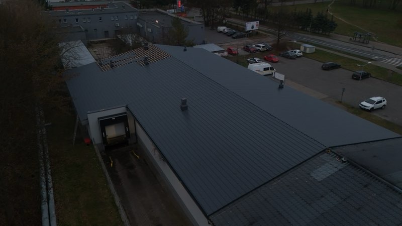 Widok z góry na budynek z szarym dachem z blachy trapezowej. Część dachu w trakcie wymiany, widoczne elementy konstrukcji. Obok parking i budynki.