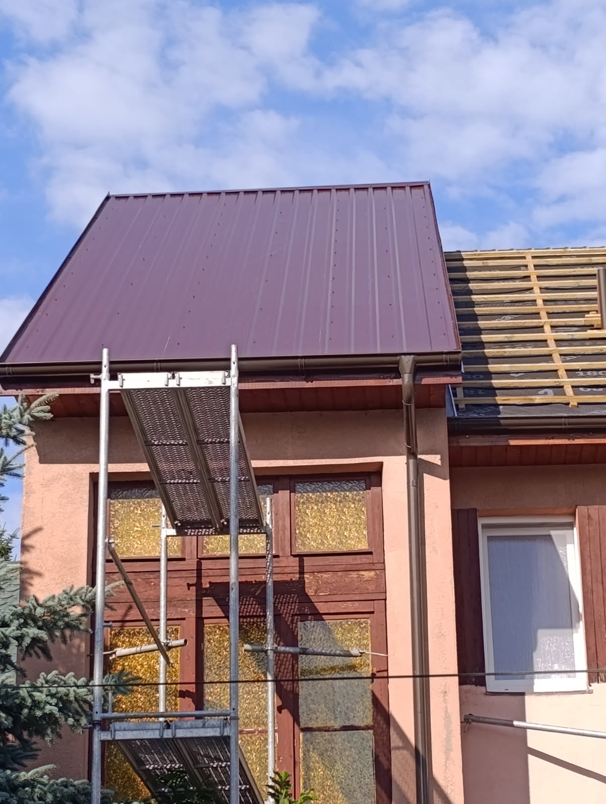 Częściowo pokryty, bordowy dach z blachy trapezowej na budynku. Widoczne rusztowanie i fragment dachu z łatami. Błękitne niebo w tle.