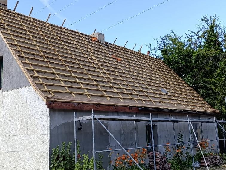 Dom w trakcie krycia dachu: widoczne łaty, membrana dachowa, częściowo ułożona dachówka. Na dole rusztowanie, kwiaty i stos dachówek. Praca w toku.