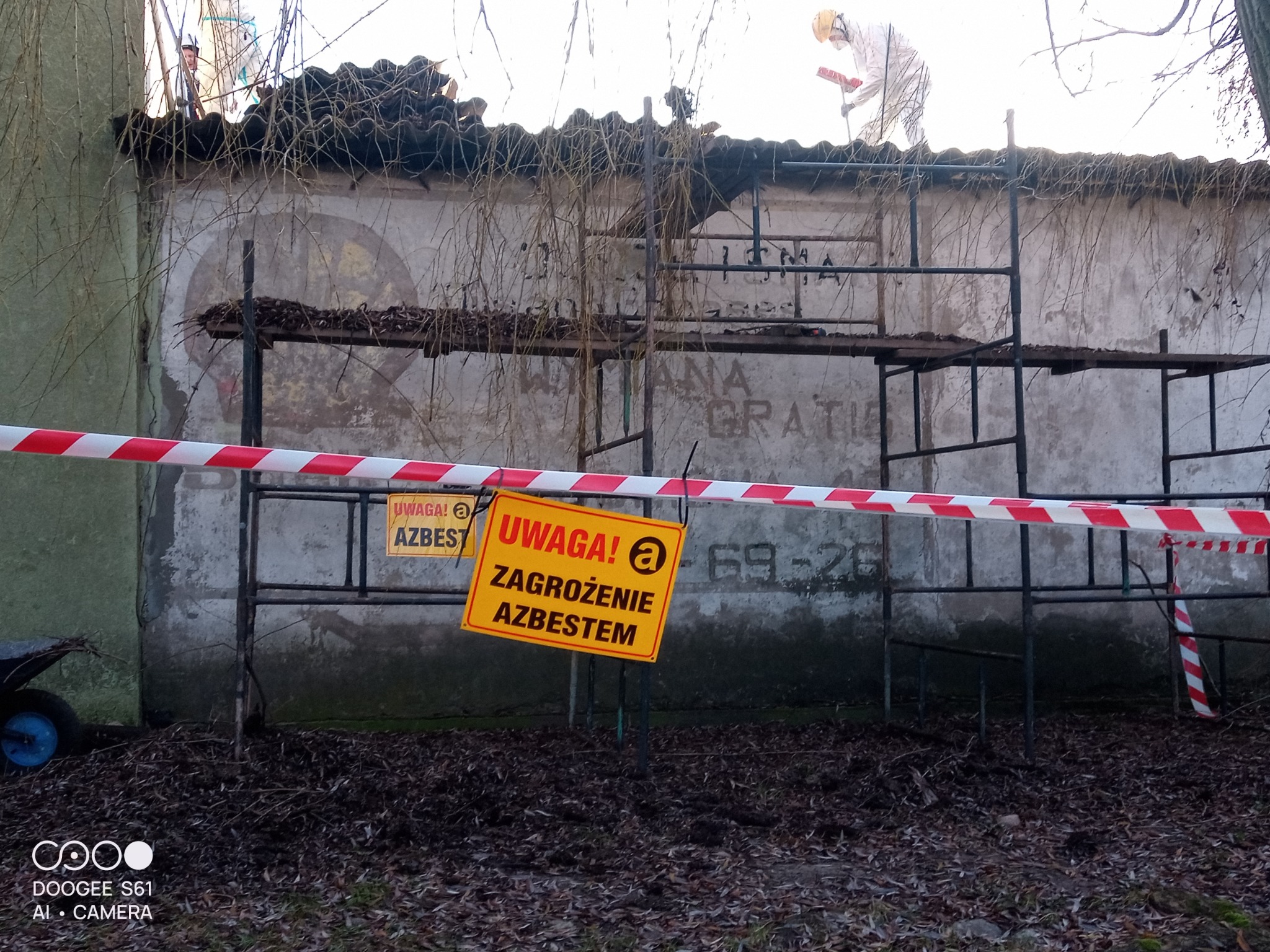 Demontaż dachu z azbestem: pracownik w kombinezonie na rusztowaniu, tablice ostrzegawcze i taśma zabezpieczająca teren. Widoczne stare napisy na ścianie budynku.