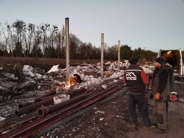 Pracownik spawa metalowe elementy konstrukcyjne na placu rozbiórki, w tle sterta gruzu i stojące słupy, dwóch mężczyzn w odzieży roboczej obserwuje prace, na plecach jednego z nich widoczne logo...
