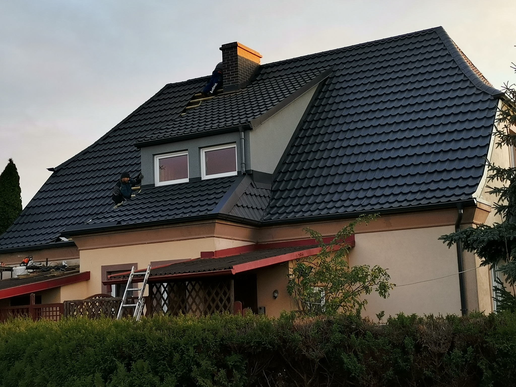 Pracownik na dachu domu jednorodzinnego z ciemną dachówką, w trakcie prac przy kominie; widoczne elementy konstrukcji dachu i narzędzia.