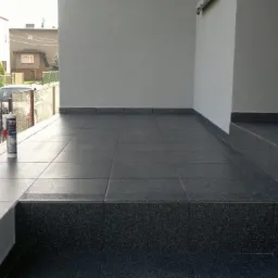 Wykończony taras z ciemnoszarą, kwadratową glazurą, widoczna tuba kleju Tytan na tle metalowego ogrodzenia i zaparkowanego samochodu.