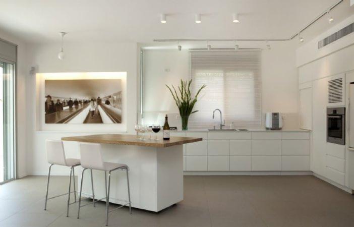 Jasne, minimalistyczne wnętrze kuchni z wyspą, wysokimi krzesłami barowymi, białymi szafkami i blatem z jasnego drewna; na ścianie czarno-biała fotografia, a na blacie kieliszki i butelka wina.