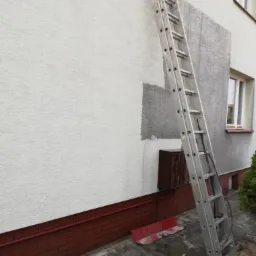 Fragment ocieplonej ściany budynku z widocznymi łatami szarego tynku, aluminiowa drabina oparta o ścianę, widoczny fragment elewacji z cegły klinkierowej.