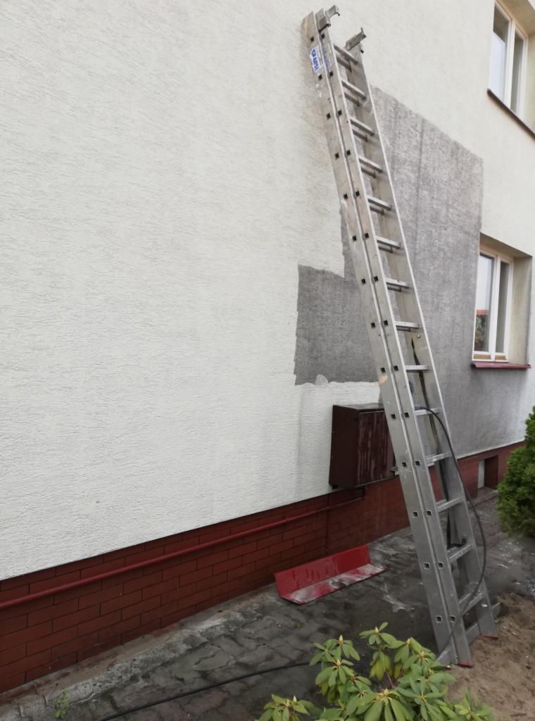 Fragment ocieplonej ściany budynku z widocznymi łatami szarego tynku, aluminiowa drabina oparta o ścianę, widoczny fragment elewacji z cegły klinkierowej.