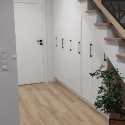 Białe szafy w zabudowie pod schodami z drewnianymi stopniami i metalową balustradą o geometrycznym wzorze, uzupełnione rośliną doniczkową, na podłodze panele imitujące jasne drewno.