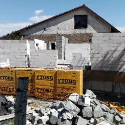 Wznoszenie ścian domu z bloczków Ytong, widoczne opakowania z bloczkami, fragment konstrukcji dachu i pracownik w tle.