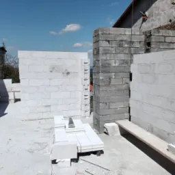 Wznoszenie ścian domu z bloczków betonowych i gazobetonowych, widoczne narzędzia murarskie i fragmenty konstrukcji dachu.