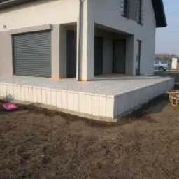 Nowoczesny taras z jasnych, prostokątnych płyt, z widocznym fragmentem elewacji domu w tle. Minimalistyczny design, surowe wykończenie.