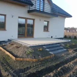 Jasny taras z kostki brukowej przy domu, obrzeża z ciemnej kostki, schody z szarej kostki, ziemia przygotowana pod nasadzenia roślin. Widok z perspektywy ogrodu.