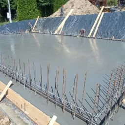 Zalany betonowy fundament z wystającymi prętami zbrojeniowymi, drewnianym szalunkiem i folią ochronną na zboczu w tle, dwóch pracowników w oddali.