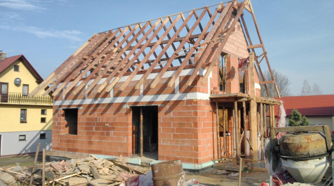 Budowa domu z czerwonej cegły: surowy stan z drewnianą konstrukcją dachu, widoczna betoniarka i gruz budowlany. Słoneczny dzień.