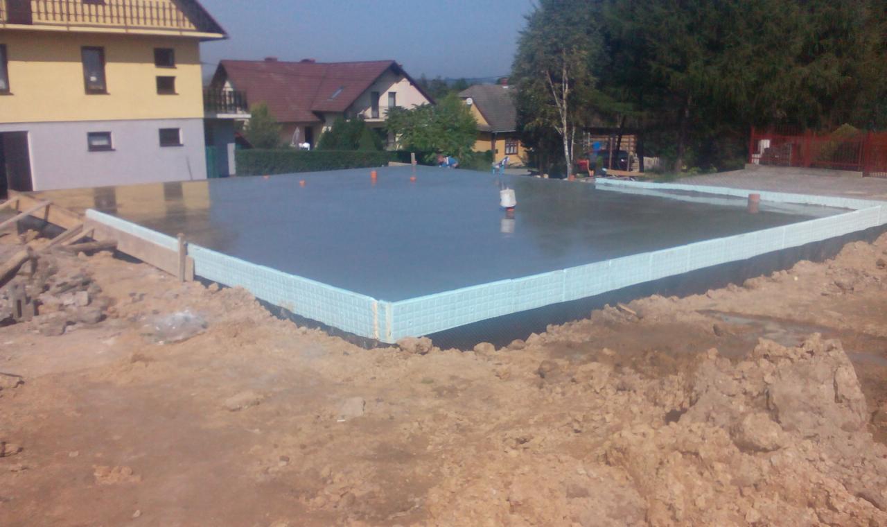 Wylany, gładki fundament z izolacją pionową z niebieskiego styropianu. W tle domy i drzewa. Widok z boku, w słoneczny dzień.
