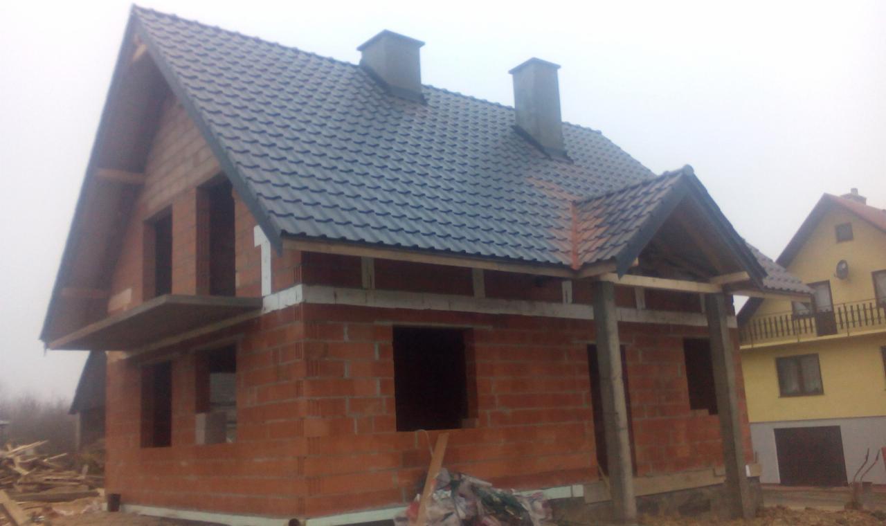 Surowy stan domu murowanego z ciemną dachówką i dwoma kominami, widoczne otwory okienne i drzwiowe, w tle inny budynek. Etap budowy przed ociepleniem.