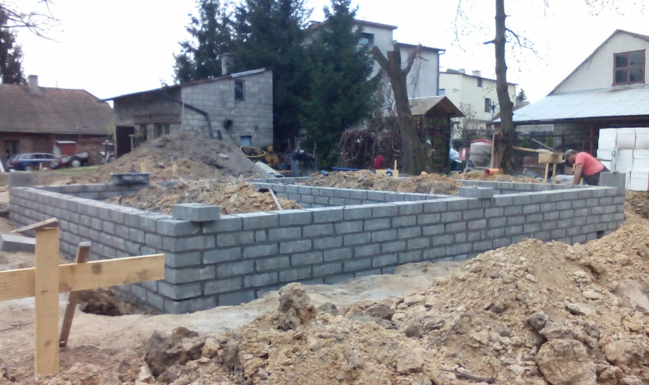 Wznoszenie fundamentów z szarej cegły na placu budowy. Widoczny fragment drewnianej konstrukcji szalunkowej i pracownik w czerwonej koszulce. Tło: zabudowa mieszkalna.