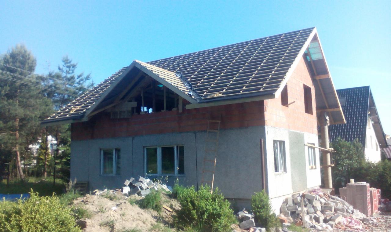 Budowa domu z keramzytu: surowy stan zadaszenia, widoczne pustaki, drewniana konstrukcja dachu i rusztowanie. Na pierwszym planie gruz i zieleń.