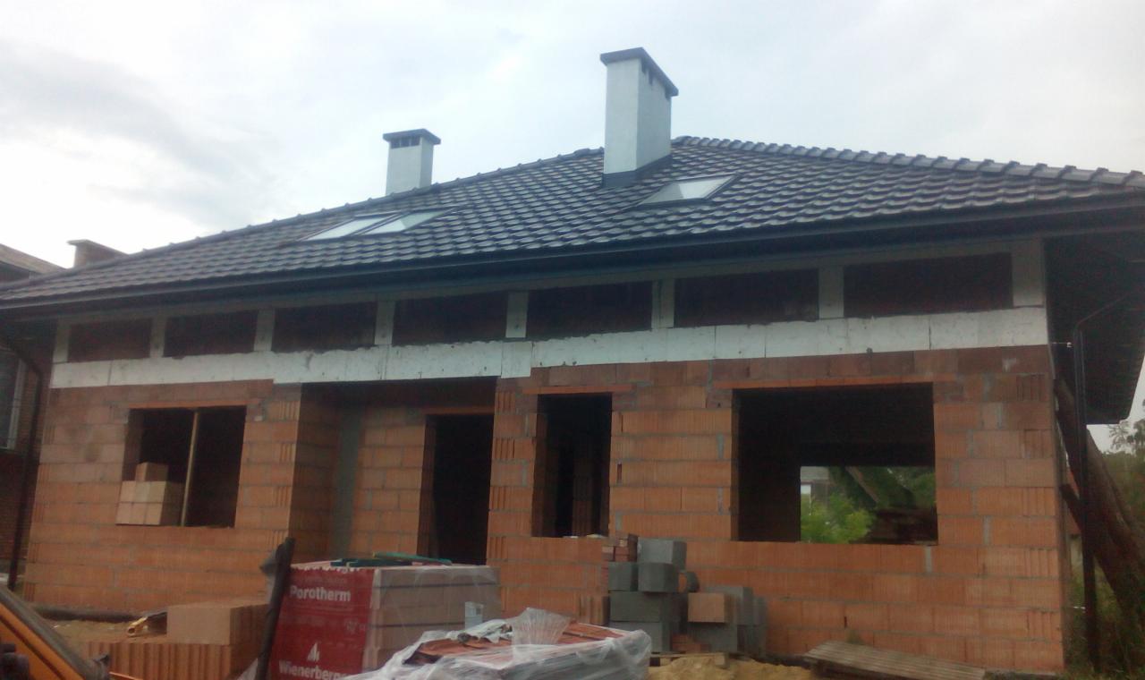 Niedokończony budynek z czerwonej cegły z widocznymi otworami okiennymi i drzwiowymi, częściowo ocieplony, z ciemnym dachem i dwoma kominami, palety cegieł Porotherm na pierwszym planie.