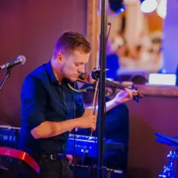 Młody mężczyzna grający na skrzypcach elektrycznych podczas występu zespołu muzycznego na tle klawiszy Kurzweil i Behringer, oświetlony światłami punktowymi.