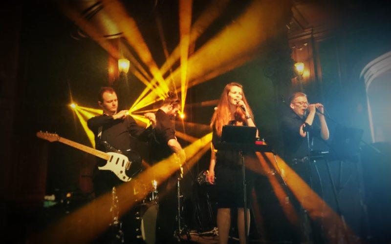 Zespół muzyczny Ars Band w trakcie występu na scenie oświetlonej reflektorami, wokalistka śpiewa do mikrofonu, gitarzysta gra na gitarze elektrycznej, a klawiszowiec gra na keyboardzie.