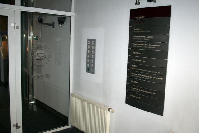 Wejście do budynku z zainstalowanym panelem domofonowym na ścianie oraz tablicą informacyjną z listą firm. Widoczny grzejnik.
