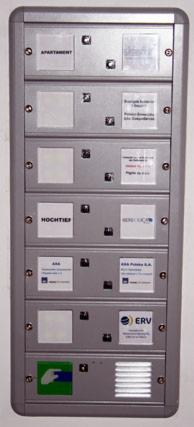Szara skrzynka pocztowa z przyciskami i miejscami na nazwiska lokatorów, zamontowana na ścianie. Widoczne logotypy firm i nazwy mieszkań.
